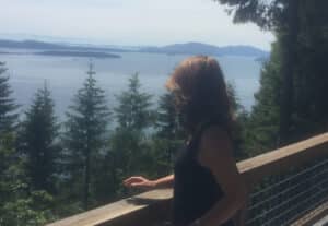 Woman standing on a wooden overlook with a railing, facing a montane lake and distant hills, trees in the foreground.